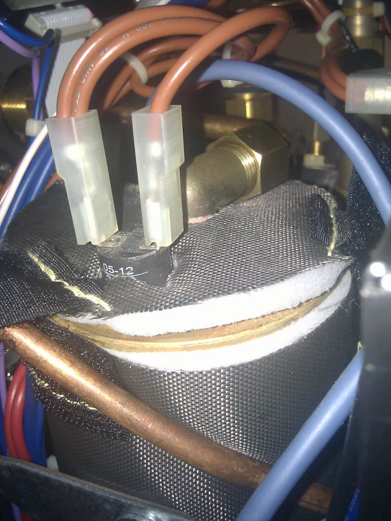 Close-up of electrical wires and connectors attached to a metal cylinder, possibly part of a machine or appliance, with various copper pipes and cables visible in the background—signs of limescale build-up are present. © 2025 1st-line.com