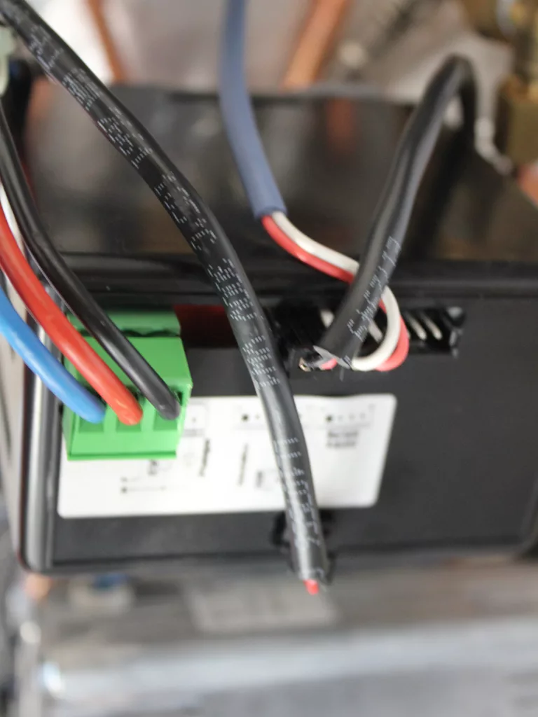 Close-up of electrical wires—black, red, blue, and white—connected to a black device with a green terminal block and a white label showing text and diagrams, commonly seen during PID troubleshooting or when resolving PID issues. © 2025 1st-line.com