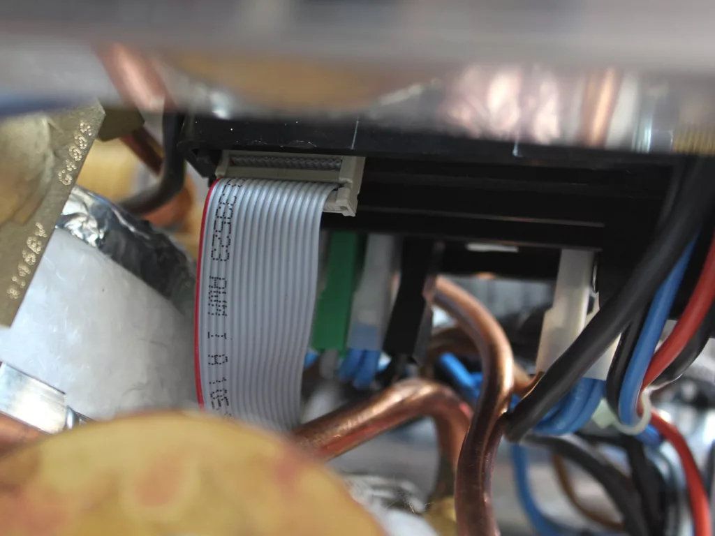 Close-up of electronic components and wiring, featuring a gray ribbon cable connected to a black connector, surrounded by copper pipes and various colored wires—ideal for troubleshooting PID display issues. © 2025 1st-line.com