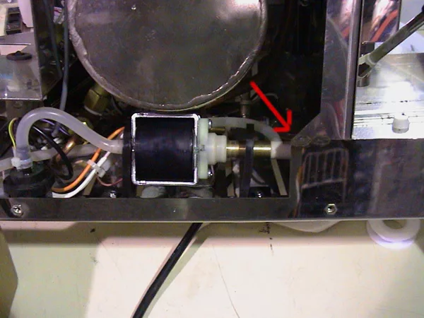 Close-up of the inner components of a machine, featuring a solenoid valve connected to clear tubing, ideal for valve repair. A red arrow points to a white plastic fitting on the right. Various wires and metal parts are visible. © 2025 1st-line.com
