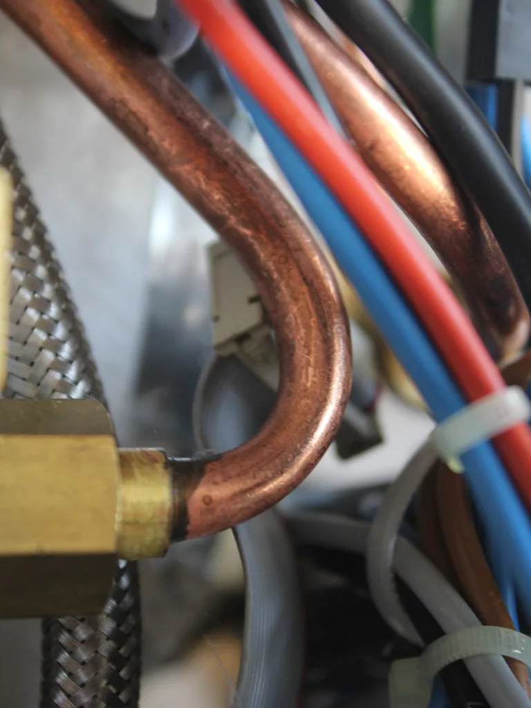 Close-up of various colorful wires, copper pipes, a braided metal hose, and a PID display inside a machine, showing interconnected mechanical and electrical components. © 2025 1st-line.com