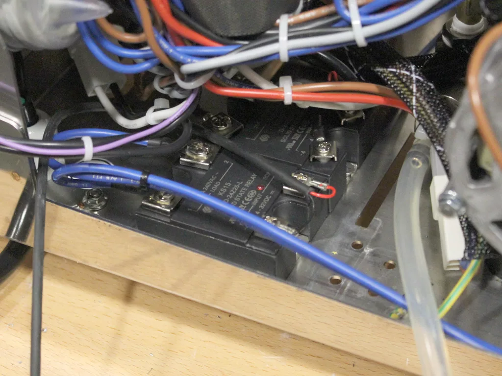 Close-up view of electrical components and wiring inside a VBM Dual Boiler machine, featuring black relays, blue and brown wires, zip ties, and a wooden base. © 2025 1st-line.com
