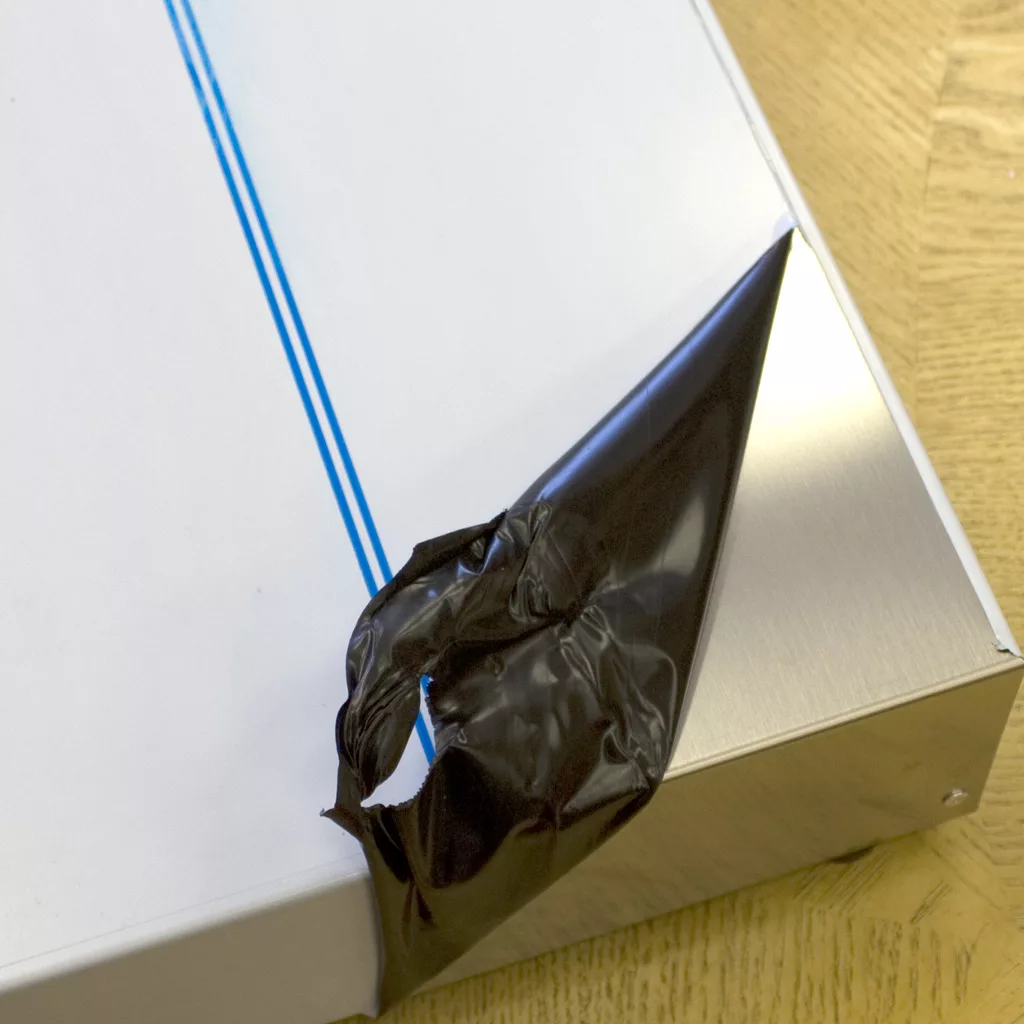 A close-up of the Ascaso I.2260 2 Drawer Base metal panel, with its white protective film being peeled to reveal a shiny surface beneath; two blue lines are visible as it rests on wood. © 2025 1st-line.com