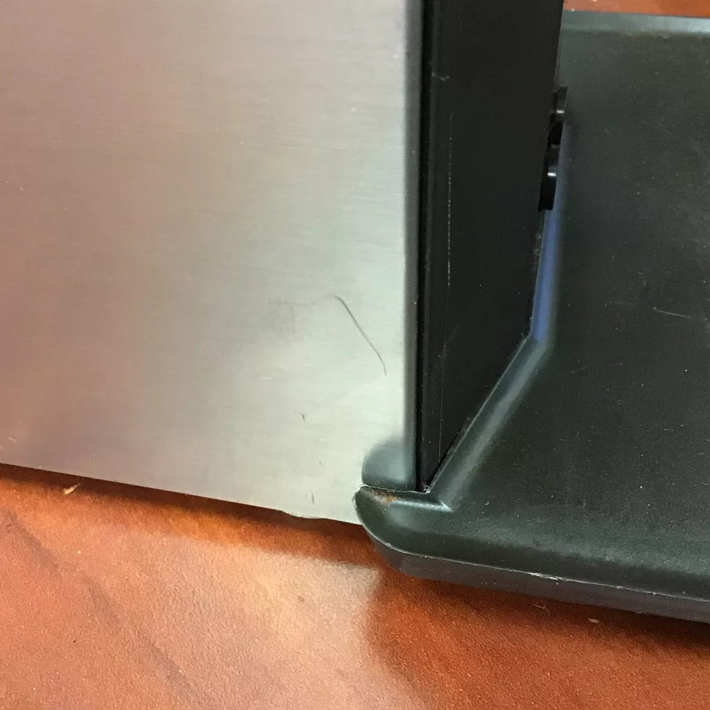 A close-up shows the corner where a SOLD Lelit PL71 William Espresso Coffee Grinder (returned & refurbished) meets its black plastic base on a wooden surface, highlighting a slight gap between the two materials. © 2025 1st-line.com