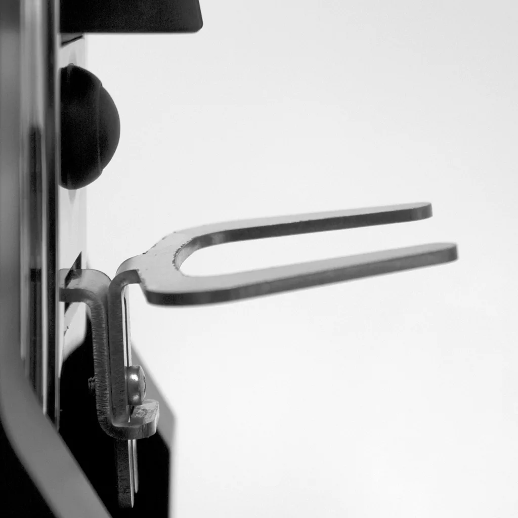 A close-up side view of a metal tuning fork clamped to a vertical black structure, evoking the sleek design of the Eureka Mignon Specialita Espresso Coffee Grinder, set against a plain light background. © 2025 1st-line.com
