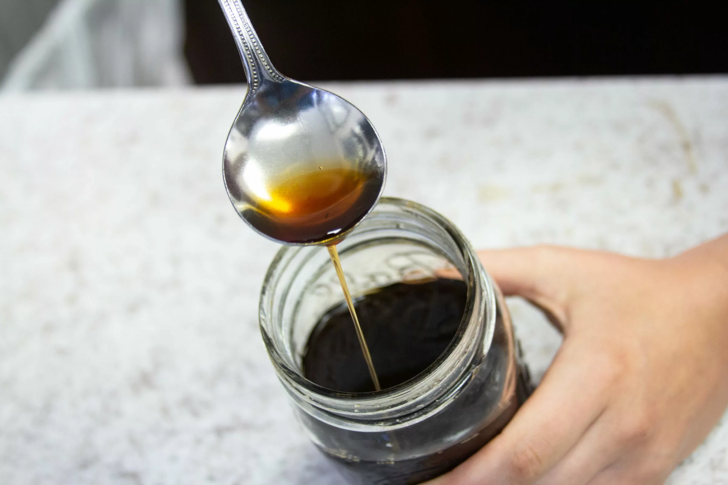 A hand holds a jar filled with dark cascara liquid while a spoon lifts some above the jar, highlighting what is cascara against a blurred background. © 2025 1st-line.com