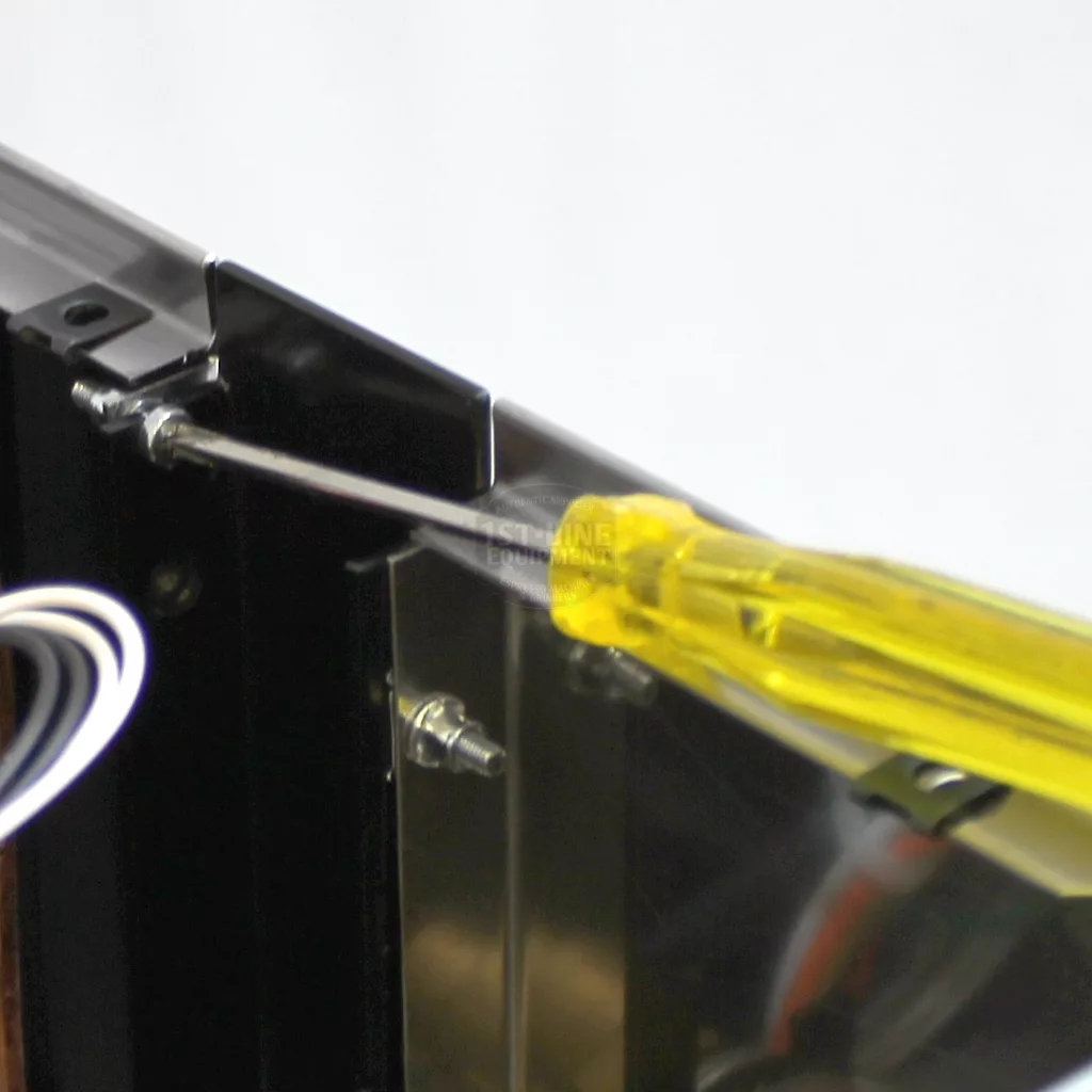 A yellow-handled screwdriver is being used to access a screw inside a black metal frame, likely while accessing the Rancilio Silvia espresso machine, with some white wires visible to the side. © 2025 1st-line.com