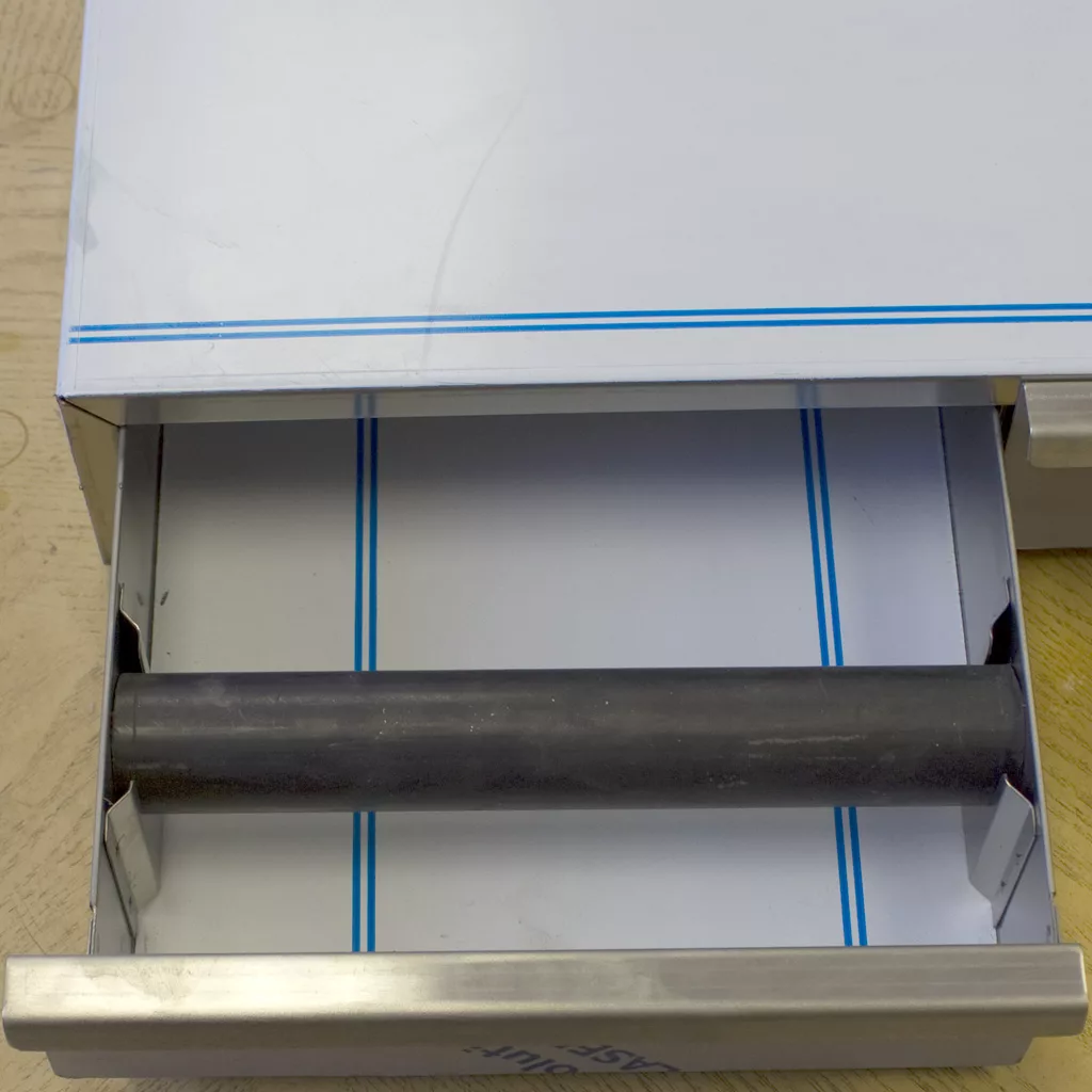 An open metal drawer from the Ascaso I.2260 2 Drawer Base reveals a cylindrical rod inside, possibly a component of industrial equipment, sitting on a light wooden surface. © 2025 1st-line.com