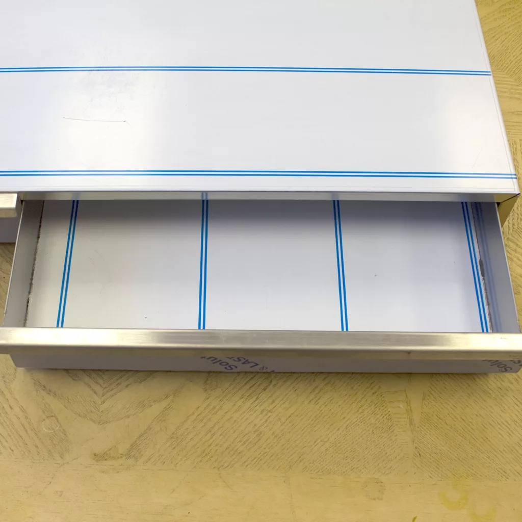 The Ascaso I.2260 2 Drawer Base features partially open metal drawers with a white interior and blue lines, set in a light wood surface. Its empty drawers highlight a clean, modern design perfect for stylish spaces. © 2025 1st-line.com