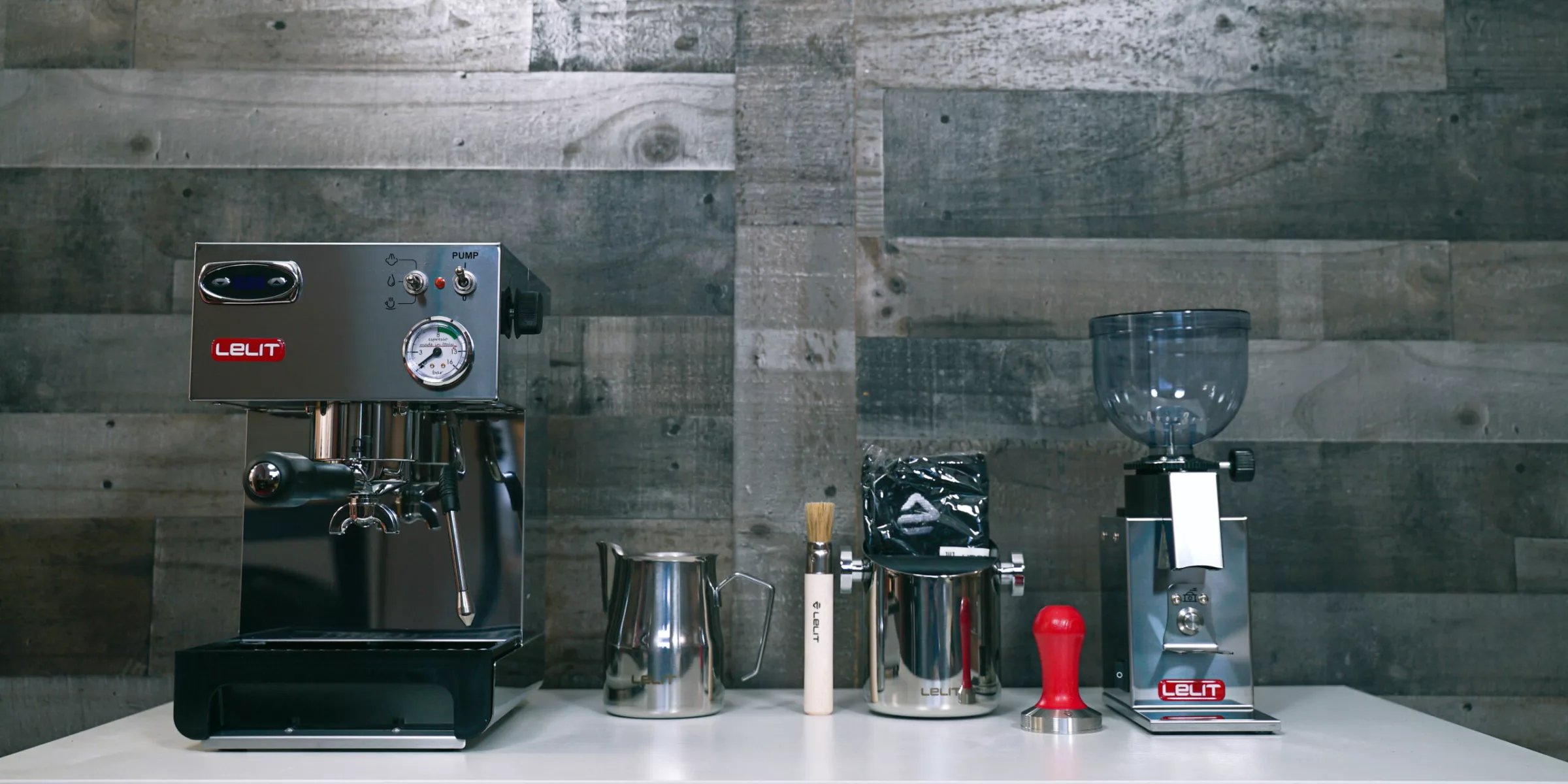 The Build My Lelit Espresso Bar includes a Lelit espresso machine, milk frothing jug, brush, tamper, coffee bag, and grinder, all neatly arranged on a white counter by a wooden plank wall. © 2025 1st-line.com