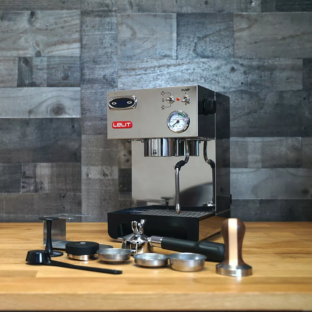 A (SOLD) SIDEWALK SALE - Lelit PL41TEM Anna Espresso Machine (returned & refurbished) sits on a wooden counter with tampers, portafilters, and accessories, set against a gray wood panel wall. © 2025 1st-line.com