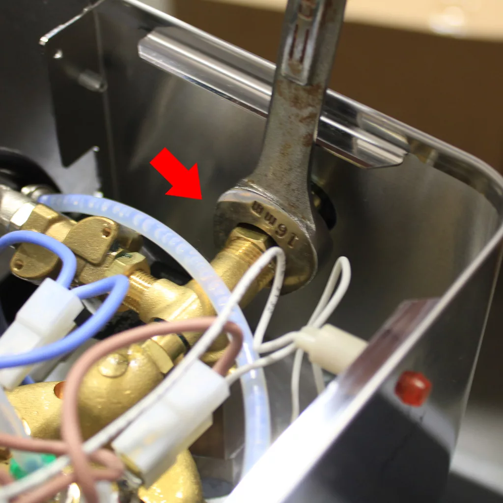 A wrench is tightening a brass fitting inside a Lelit machine, with an arrow pointing to the connection. Several wires and a transparent tube are visible nearby, illustrating a typical setup found on the Support Page. © 2025 1st-line.com