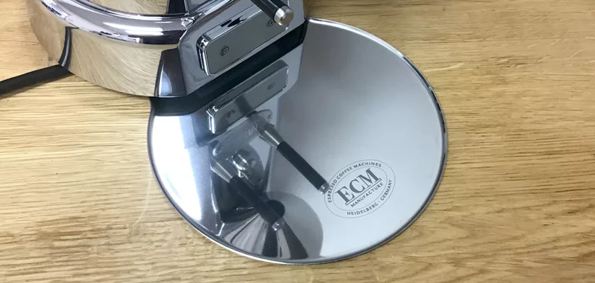 Close-up of a shiny metallic tamper with the ECM Heidelberg logo on its base, resting on a wooden surface beside a DISCONTINUED ECM V-Titan 64 Espresso Grinder. © 2025 1st-line.com