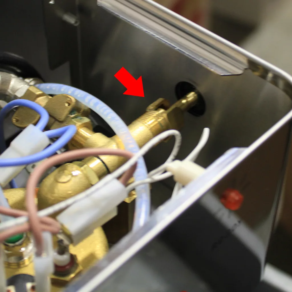 Close-up of brass and plastic components inside a Lelit machine, with wires and tubes visible. A red arrow points to a specific brass fitting on the metal wall of the enclosure—refer to our Support Page for technical support. © 2025 1st-line.com