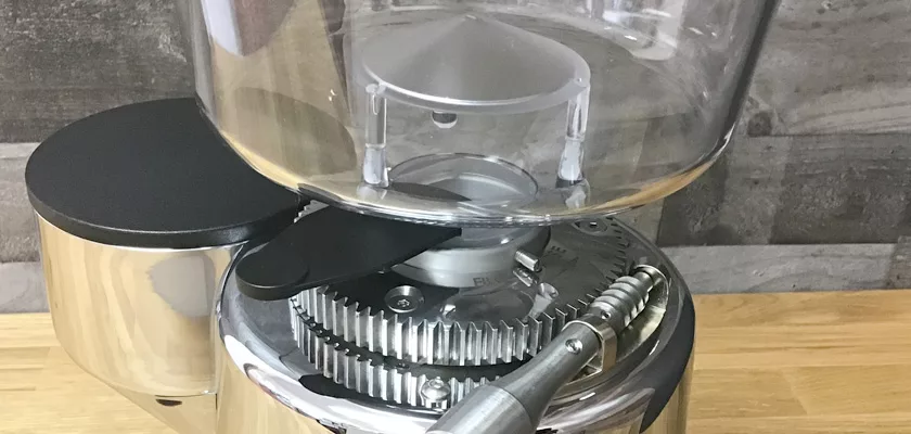 Close-up of the DISCONTINUED ECM V-Titan 64 Espresso Grinders top showcases its hopper and large exposed burrs featuring metallic and transparent parts, all set on a wooden countertop with a stone tile background. © 2025 1st-line.com