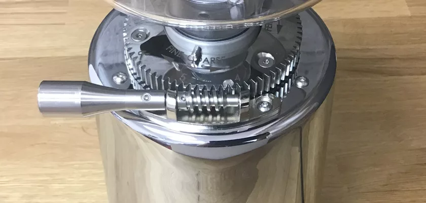 Close-up of the shiny metal DISCONTINUED ECM V-Titan 64 Espresso Grinder with hand crank and adjustable grind settings, resting on a wooden surface. This manual model is no longer available. © 2025 1st-line.com