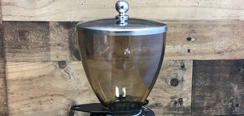 A glass coffee bean hopper with a metal lid, designed for the Elektra MSD Espresso Coffee Grinder, rests on a scale atop a rustic wooden surface. © 2025 1st-line.com