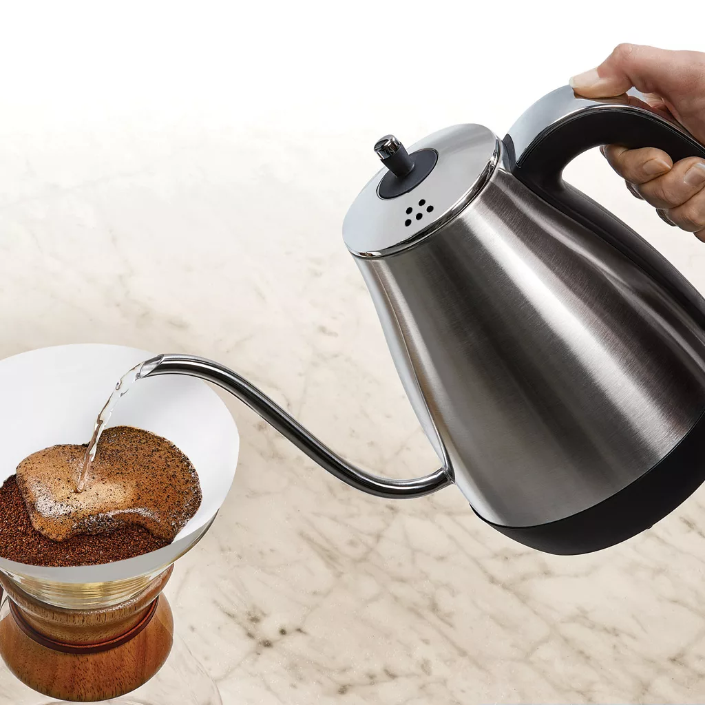 A hand pours hot water from the Capresso 290.05 Electric Pour Over Water Kettle over ground coffee in a white dripper on a wooden stand, making pour-over coffee on a marble countertop. © 2025 1st-line.com