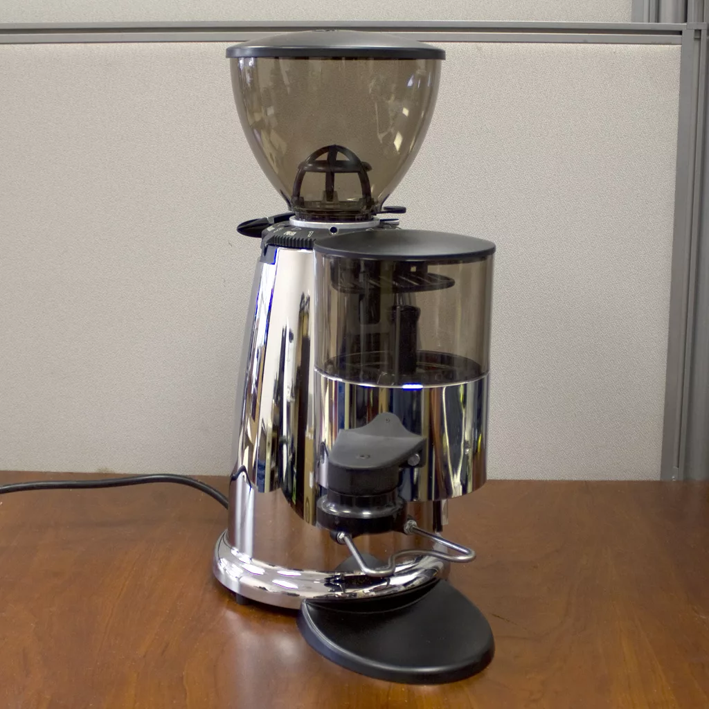 A SOLD Macap M42 Stepless Doser Espresso Coffee Grinder (demo) in silver and black sits on a wooden table against a light partition, its power cord extending from the back. © 2025 1st-line.com