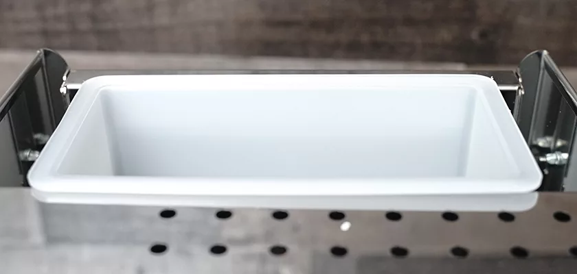 A close-up of an empty, rectangular white plastic tray inside a stainless steel container with small round holes, designed for the DISCONTINUED ECM Mechanika V Slim Espresso Machine. © 2025 1st-line.com