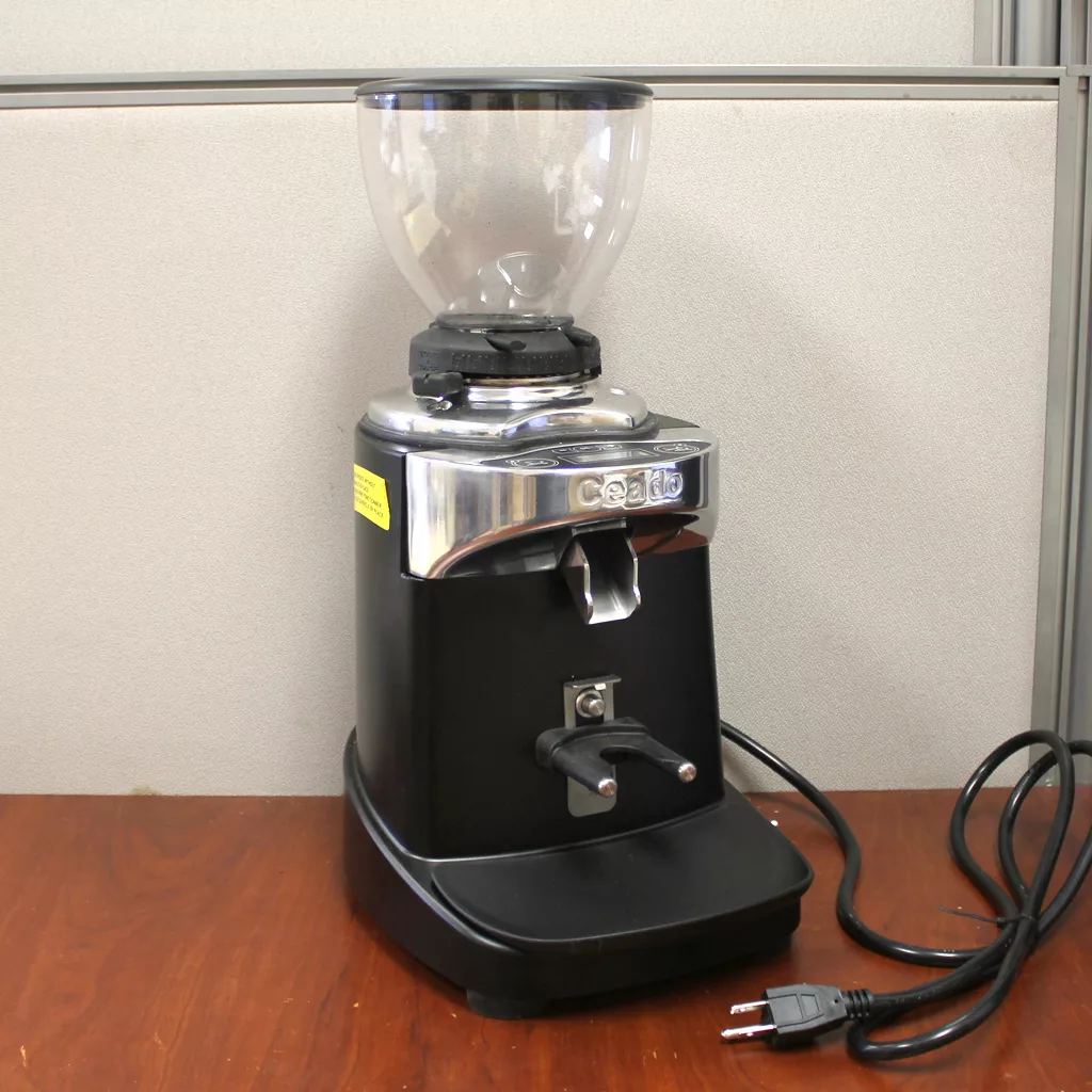 A SOLD Ceado E37J Commercial Espresso Coffee Grinder (returned & refurbished) in black and silver with a clear hopper rests on a wooden surface near a light partition, its coiled power cord positioned to the right. © 2025 1st-line.com