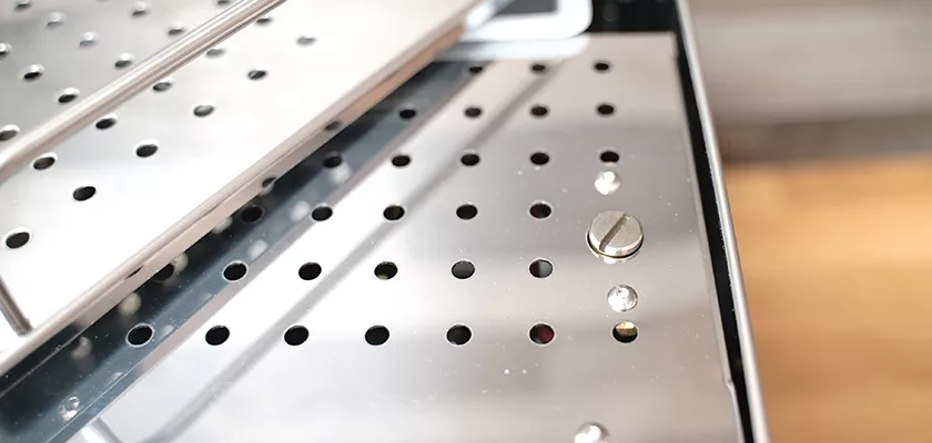 Close-up of a shiny metallic tray with round holes, a flat-head screw, and small raised bumps; likely part of the discontinued ECM Mechanika V Slim Espresso Machine. © 2025 1st-line.com