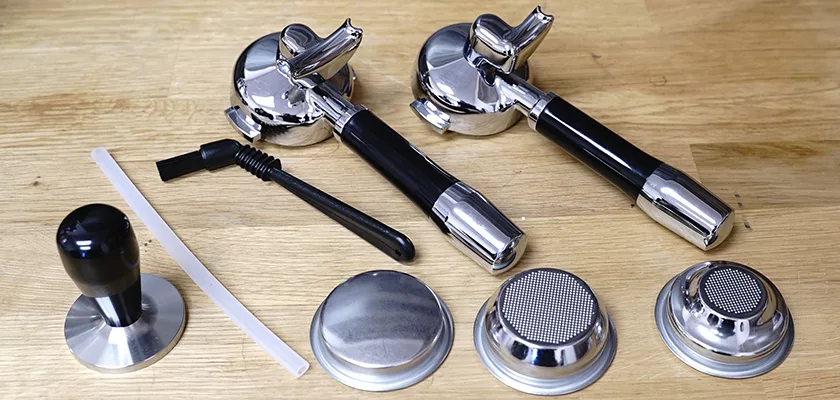 Parts and accessories for the DISCONTINUED ECM Mechanika V Slim Espresso Machine—two portafilters, a tamper, cleaning brush, plastic tool, and metal filter baskets—are neatly arranged on a wooden surface. © 2025 1st-line.com