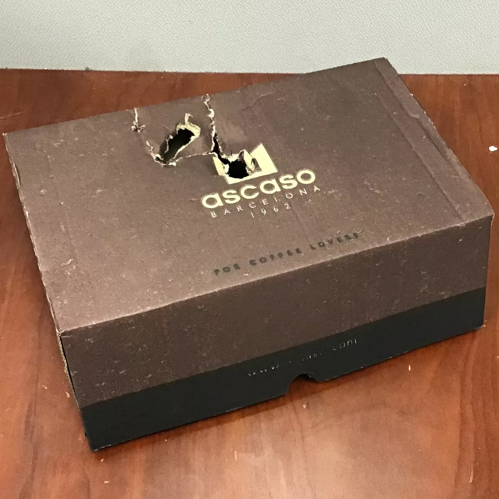 A brown Ascaso Barcelona box with gnawed holes in the lid, labeled “For Coffee Lovers,” sits on wood—perhaps holding a SOLD Ascaso Dream UP V3 Anthracite Gray PID Espresso Machine (returned & refurbished). © 2025 1st-line.com