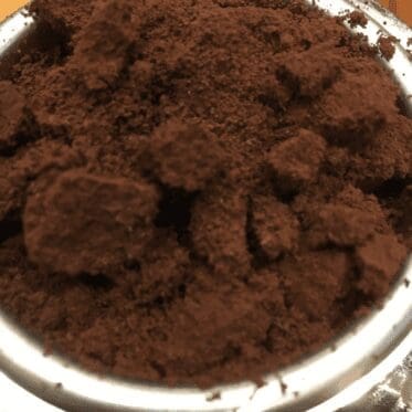 A close-up of freshly ground coffee in a metal portafilter, inviting you to consider spending a little more for quality beans. The dark brown grounds appear coarse, perfect for brewing espresso. © 2025 1st-line.com