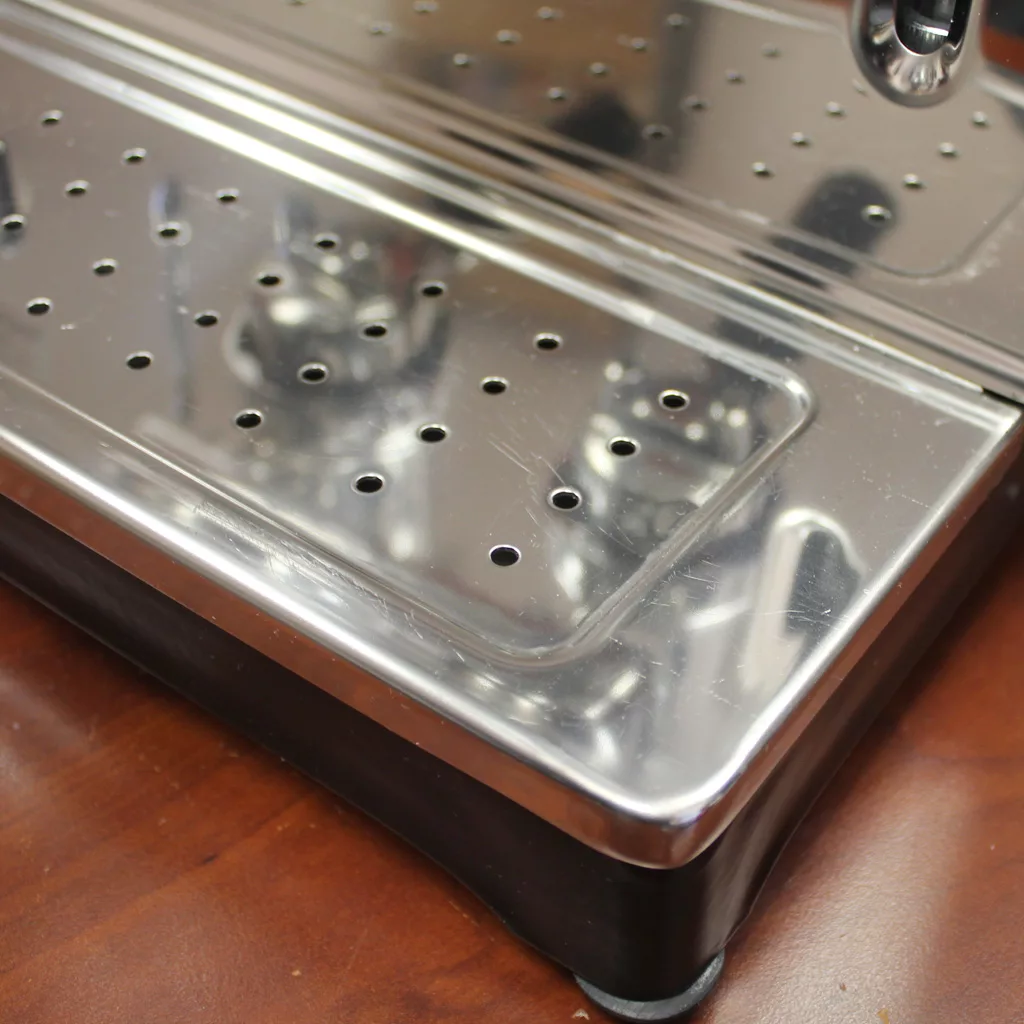 Close-up of the shiny metal drip tray with circular holes on a returned & refurbished SOLD Ponte Vecchio Lusso Espresso Machine - 2 group, chrome, resting on a wooden countertop. © 2025 1st-line.com