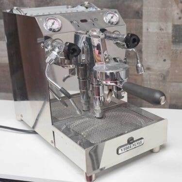 The SIDEWALK SALE - VBM Domobar Super V4 Dual Boiler Switchable Espresso Machine (USED) (E109) sits on a white countertop against a rustic wooden wall, featuring stainless steel construction, gauges, levers, and portafilters. © 2025 1st-line.com