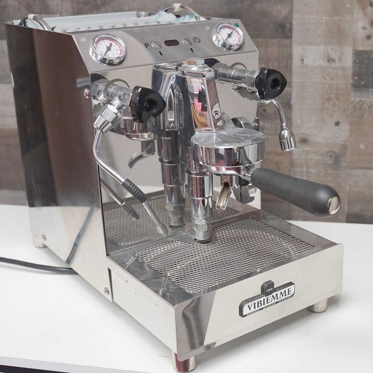 The SIDEWALK SALE - VBM Domobar Super V4 Dual Boiler Switchable Espresso Machine (USED) (E109) sits on a white countertop against a rustic wooden wall, featuring stainless steel construction, gauges, levers, and portafilters. © 2025 1st-line.com