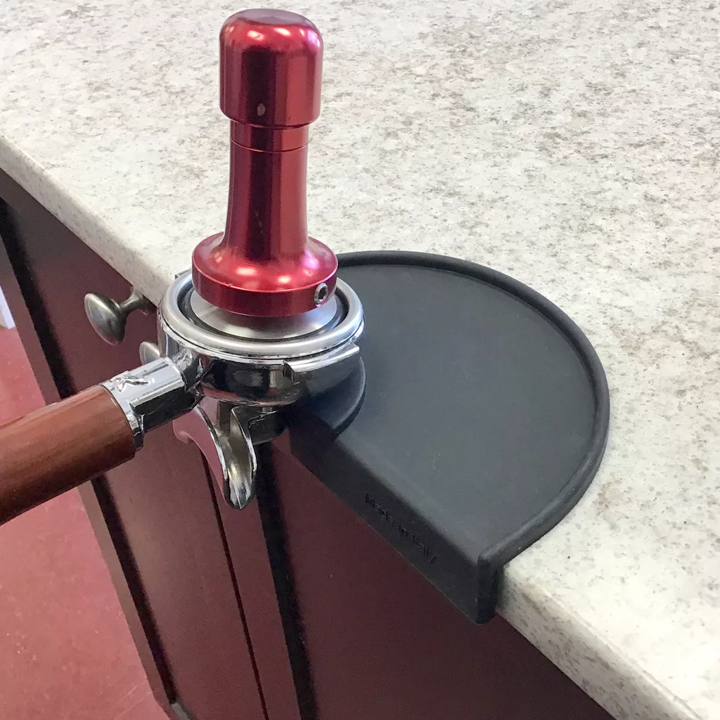A metal espresso tamper rests on a portafilter, which is positioned on a 1st-line Countertop Edge Tamping Mat at the edge of a countertop. © 2025 1st-line.com