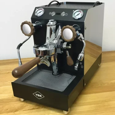 A VBM espresso machine featuring the Walnut Wood Kit—knobs and portafilter handle—sits ready on a wooden countertop, combining elegant walnut accents with dual gauges for brewing coffee. © 2025 1st-line.com