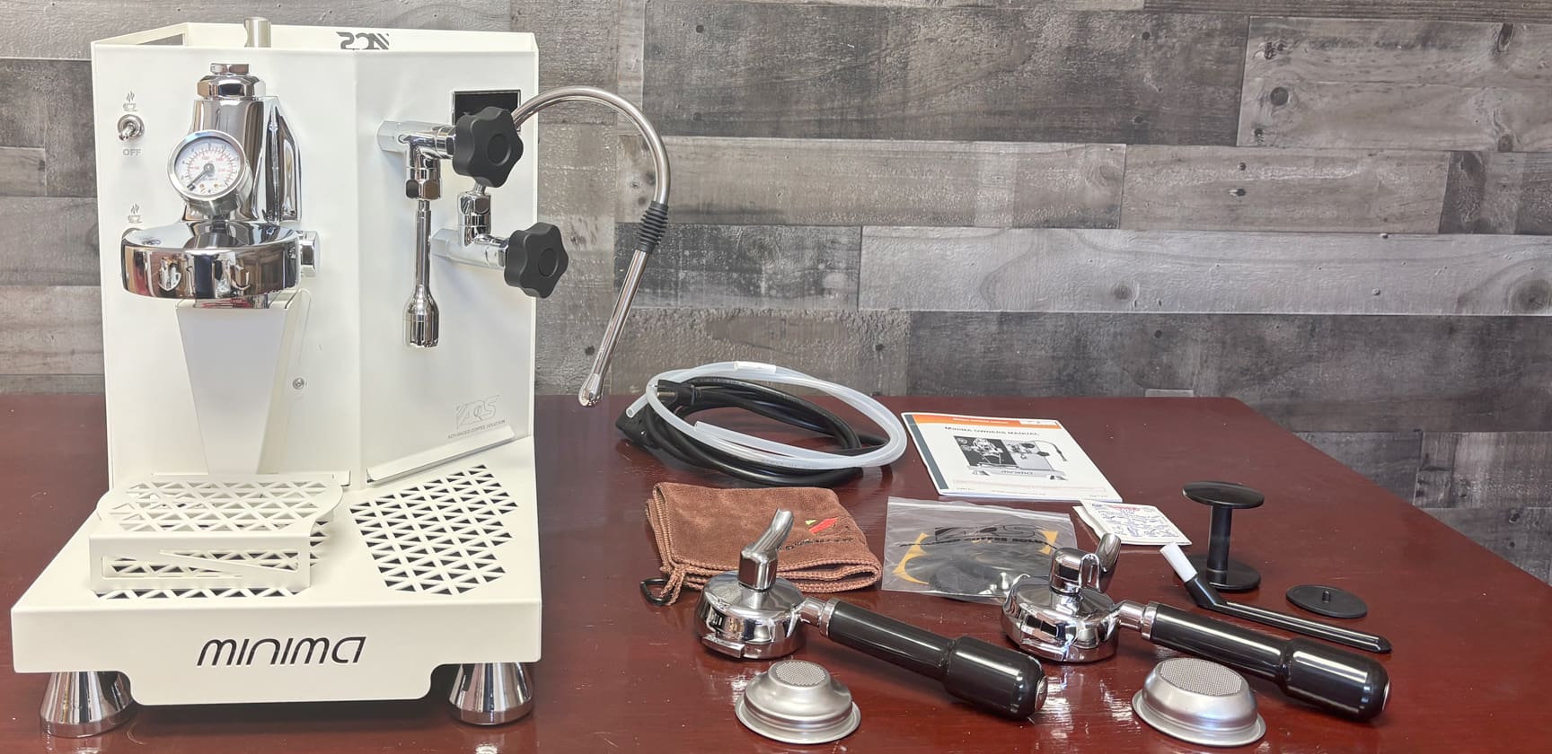 An ACS Minima Dual Boiler Espresso Machine in white rests on a wooden table with included accessories: portafilters, filters, tamper, hoses, cleaning brush, and manuals, set against gray wooden panels. © 2025 1st-line.com
