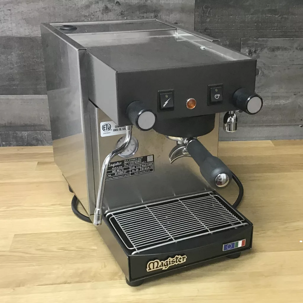 A refurbished Magister MS40 Stilo HX Tank Espresso Machine, for in-store pickup only, sits on a wooden counter with dual switches, a steam wand, and portafilter. The front displays the Magister logo and an Italian flag. © 2025 1st-line.com