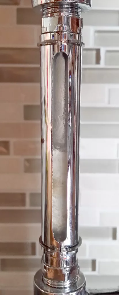 A close-up of a chrome water pipe with a cloudy sight glass revealing a buildup of white mineral deposits inside, set against a blurred tiled wall background. © 2025 1st-line.com