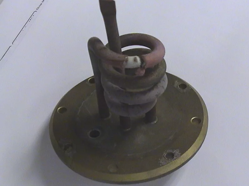 A metal plate with several holes and attached tubes, including a coiled tube, is resting on a white surface. This object appears to be a heating element from an Elektra Microcasa Semiautomatica espresso machine. © 2025 1st-line.com