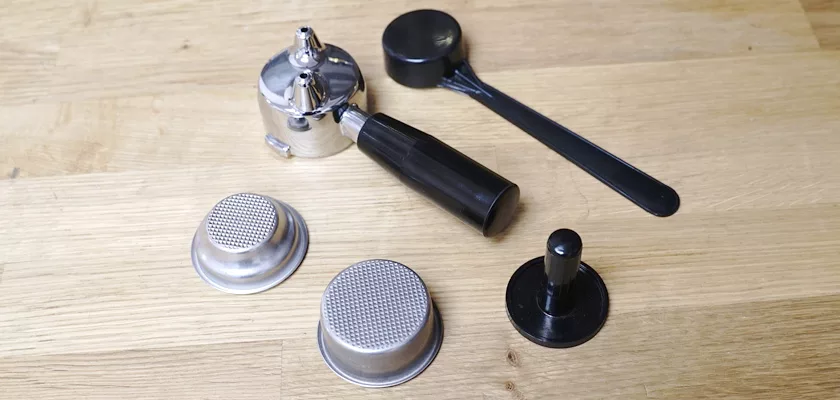 Accessories for the Elektra Microcasa a Leva Espresso Machine—a portafilter, two metal filter baskets, a black coffee scoop, and a tamper—are displayed on a light wooden surface. © 2025 1st-line.com