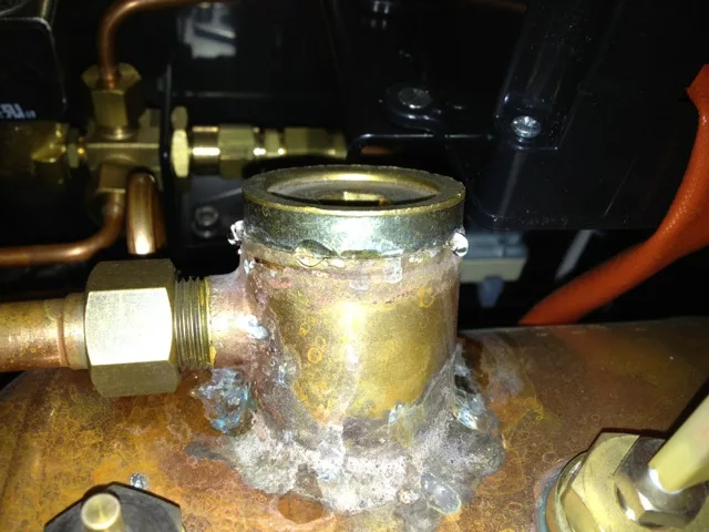Close-up of a brass pipe joint on an Elektra espresso machine with visible mineral buildup and corrosion, showing water leakage around the leaking HX access port and drops forming on the surface. © 2025 1st-line.com