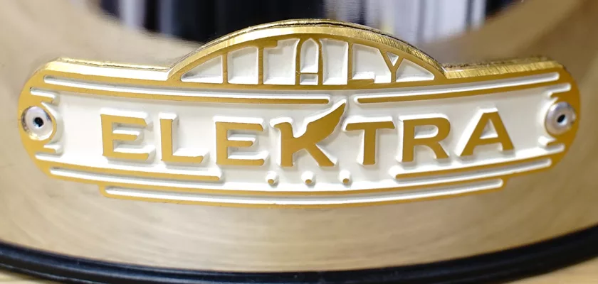 Close-up of a metallic nameplate reading ELEKTRA in gold letters with an arc and building design, attached by two screws to the reflective surface of the Elektra Microcasa a Leva Espresso Machine. © 2025 1st-line.com