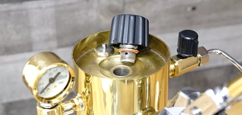 Close-up of the Elektra Microcasa a Leva Espresso Machine with a gold metal body, open threaded opening, black plastic knob, pressure gauge, and detailed components set against a gray textured background. © 2025 1st-line.com