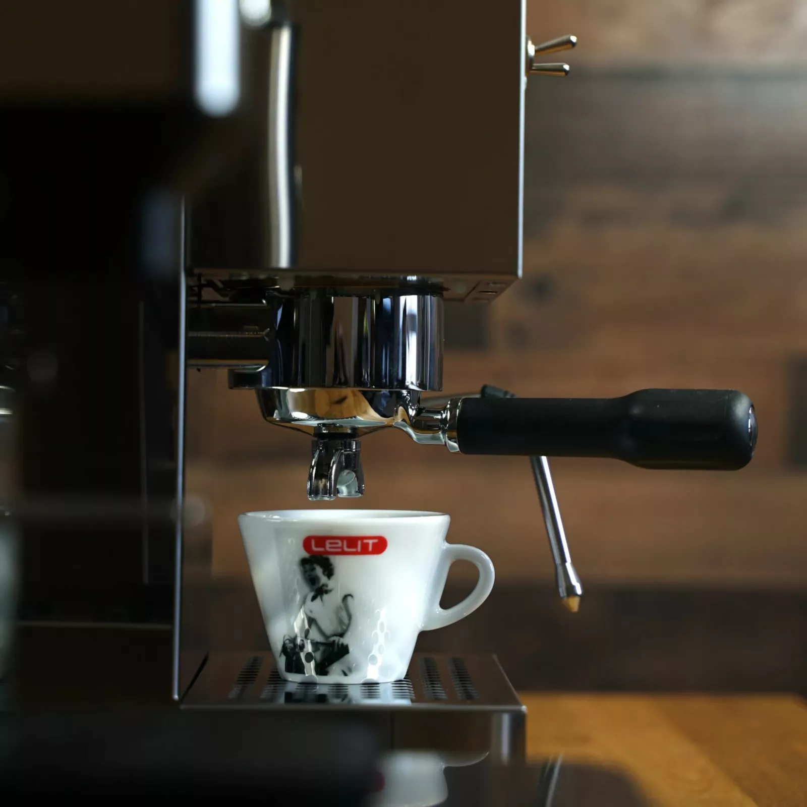 A close-up of a DISCONTINUED Lelit Glenda PL41PLUST PID Espresso & Cappuccino Machine brewing coffee into a white LeLit cup on a wooden counter, with a blurred brown background. © 2025 1st-line.com