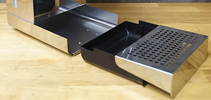 A close-up of the DISCONTINUED Lelit Glenda PL41PLUST PID Espresso & Cappuccino Machine’s drip tray, partially pulled out on a wooden countertop. The tray has a black plastic base and a metal cover with round holes. © 2025 1st-line.com