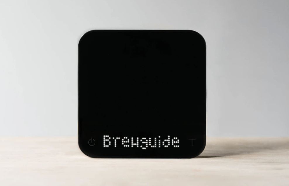 A DISCONTINUED Acaia Pearl Model S Professional-Grade Smart Scale in black, featuring a square design, rests on a light wooden surface. The display shows Brewguide in white dot matrix text against a simple, light background. © 2025 1st-line.com