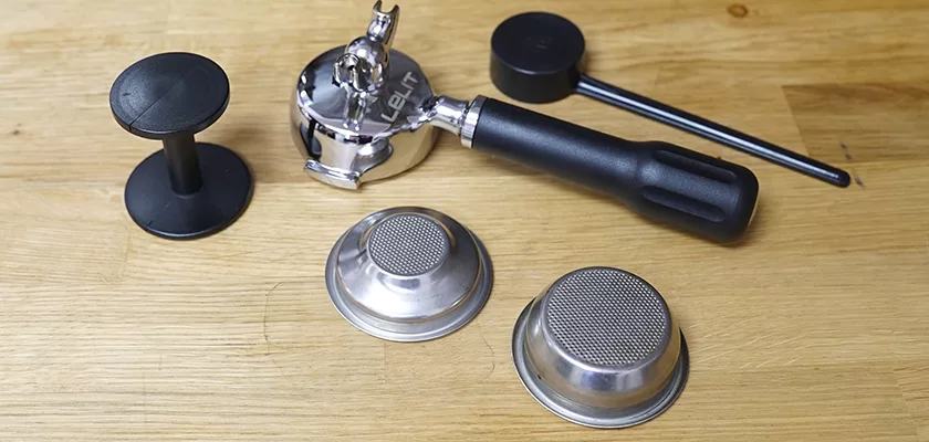 A portafilter with handle, two metal filter baskets, a black tamper, and a black dosing tool are arranged on wood—ideal accessories to enhance your DISCONTINUED Lelit Glenda PL41PLUST PID Espresso & Cappuccino Machine setup. © 2025 1st-line.com