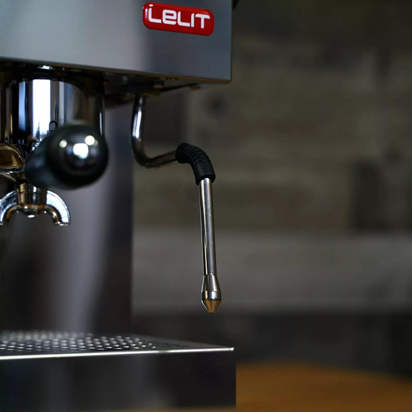 Close-up of the discontinued Lelit Glenda PL41PLUST PID Espresso & Cappuccino Machine, featuring a steam wand and red Lelit label against a blurred gray brick wall and wooden surface background. © 2025 1st-line.com