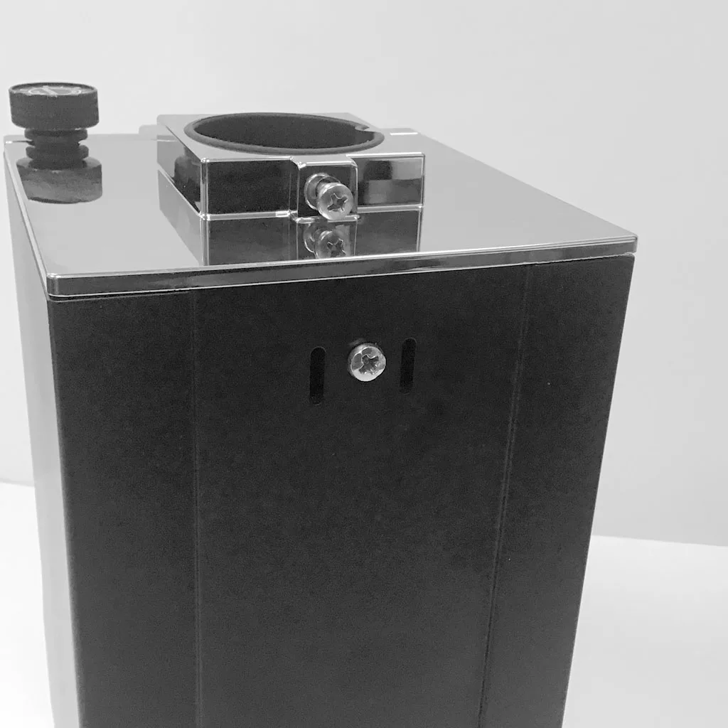 A black and silver rectangular metal device, resembling the Eureka Mignon Grinder, features a circular opening for accessing burrs and a knob on top, set against a plain white background. © 2025 1st-line.com