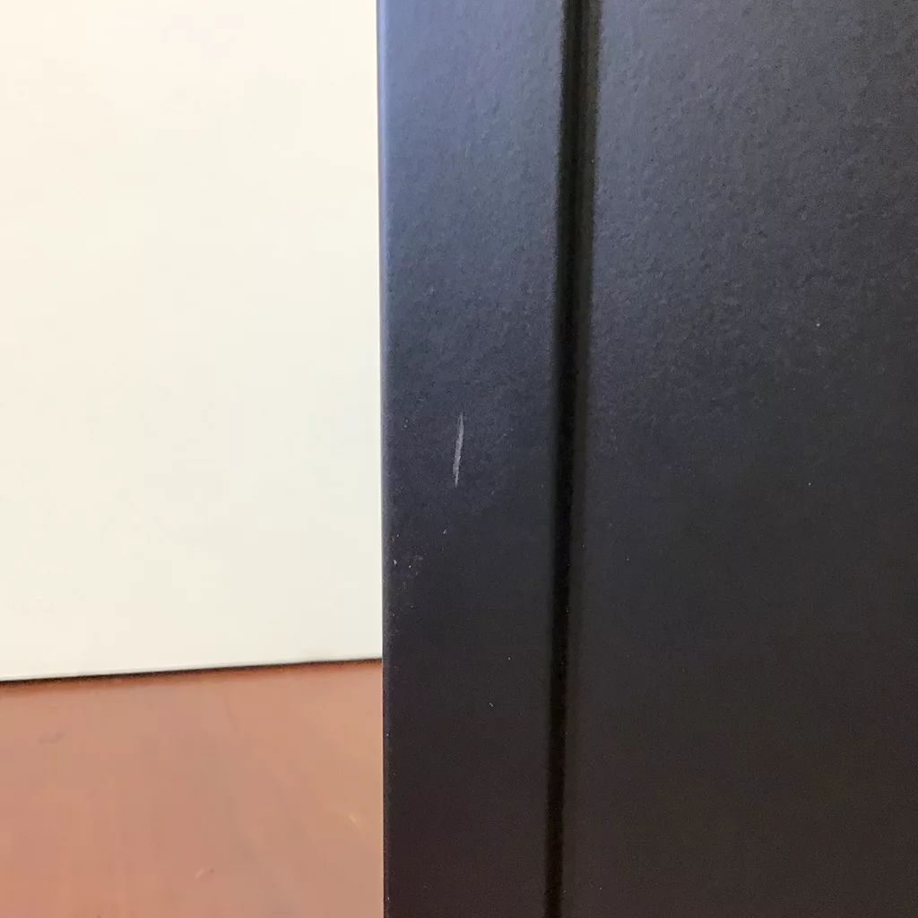 A close-up of a SOLD Eureka Mignon Specialita Stepless Doserless Espresso Coffee Grinder (returned & refurbished) highlights its black finish with a small white scratch, set on a brown floor with a white wall in the background. © 2025 1st-line.com