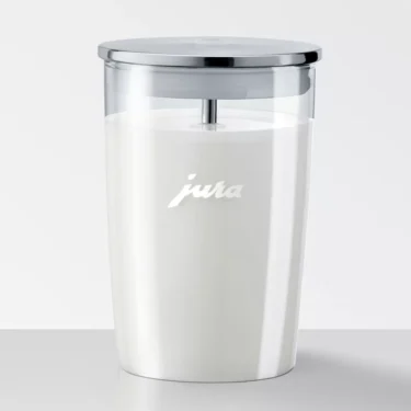 A Jura Glass Milk Container with Lid - 16.9 oz, filled with milk, features a stainless steel lid, internal metal rod, and white jura branding on the front. The container rests on a light surface against a white background. © 2025 1st-line.com